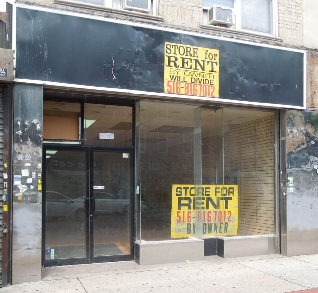 A vacant storefront with large glass windows and a black awning with faded writing. A "STORE FOR RENT" sign with contact information is prominently displayed in the window. The interior is empty with visible shelves on the right side. The sidewalk is cracked.