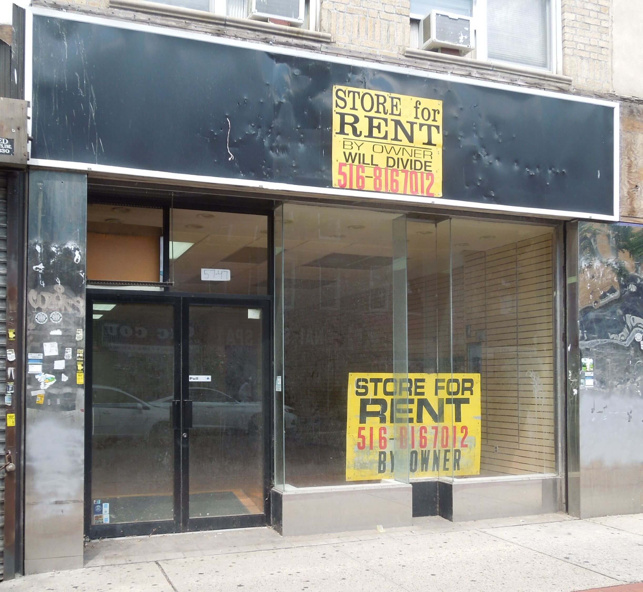 A vacant storefront with large glass windows and a black awning with faded writing. A "STORE FOR RENT" sign with contact information is prominently displayed in the window. The interior is empty with visible shelves on the right side. The sidewalk is cracked.
