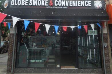 A storefront with a sign reading "Globe Smoke & Convenience" with the subtitle "full line of tobacco products." The storefront has a glass front with shelves visible inside. Red, white, and blue pennant flags are strung across the top of the entrance.