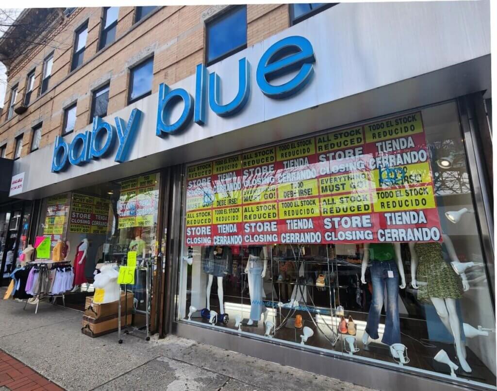 Storefront with large blue "baby blue" sign. Windows display "Store Closing" and "All Stock Reduced" signs in English and Spanish. Mannequins in trendy clothing are visible. Sale items are displayed on racks outside the entrance on the sidewalk.