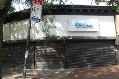 A closed storefront with a faded “Sterling Optical” sign, metal shutters down, and a “No Standing Anytime” street sign in front; trees provide shade on the sidewalk.