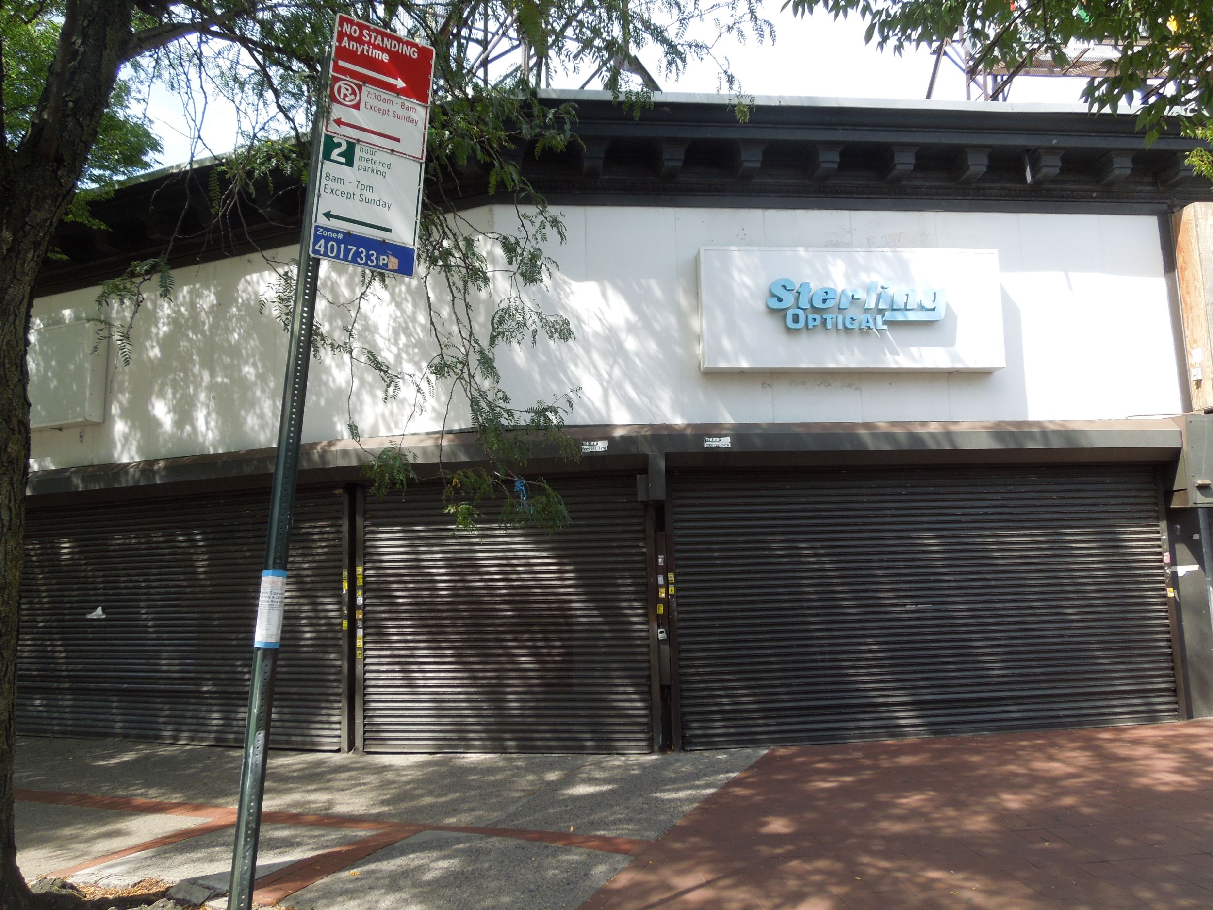 A closed storefront with a faded “Sterling Optical” sign, metal shutters down, and a “No Standing Anytime” street sign in front; trees provide shade on the sidewalk.