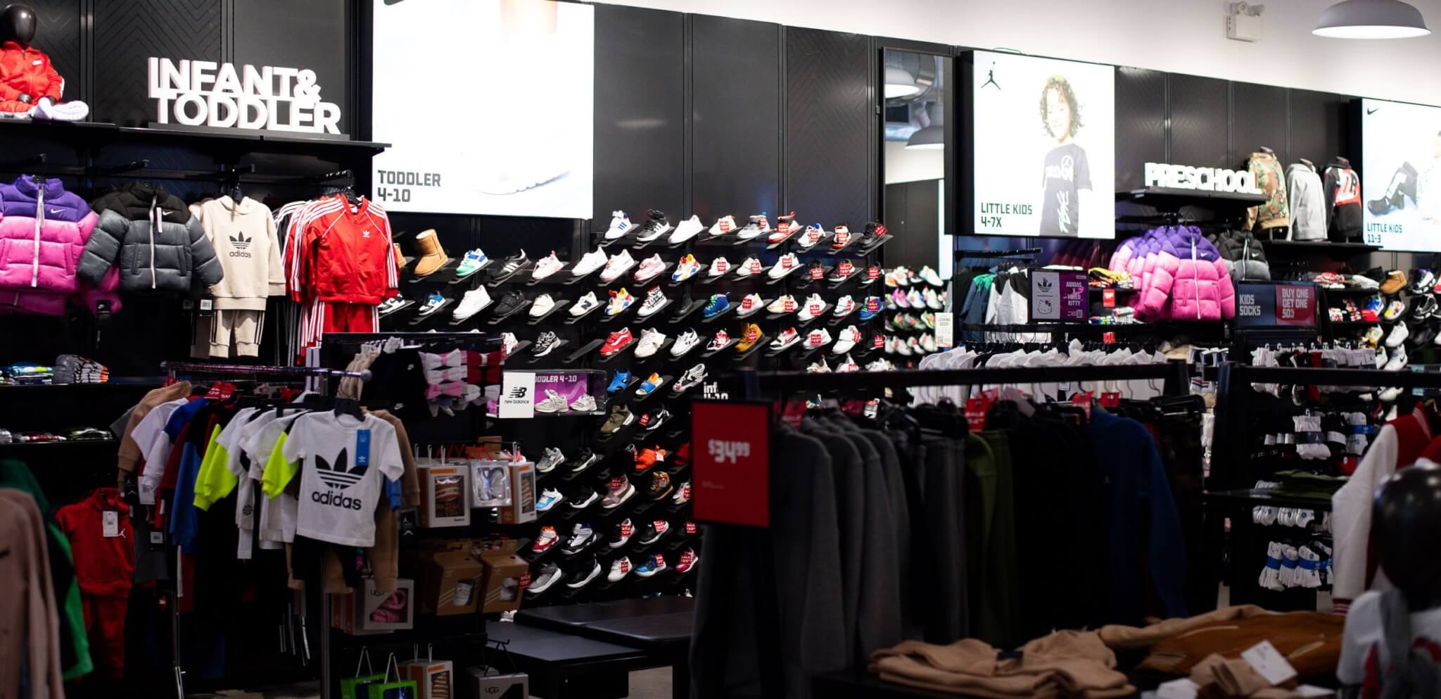 Clothing store section with racks of kids’ clothes, a wall displaying children’s shoes, and signs for “INFANT & TODDLER” and “PRESCHOOL.” Bright lighting and various colorful apparel are visible.